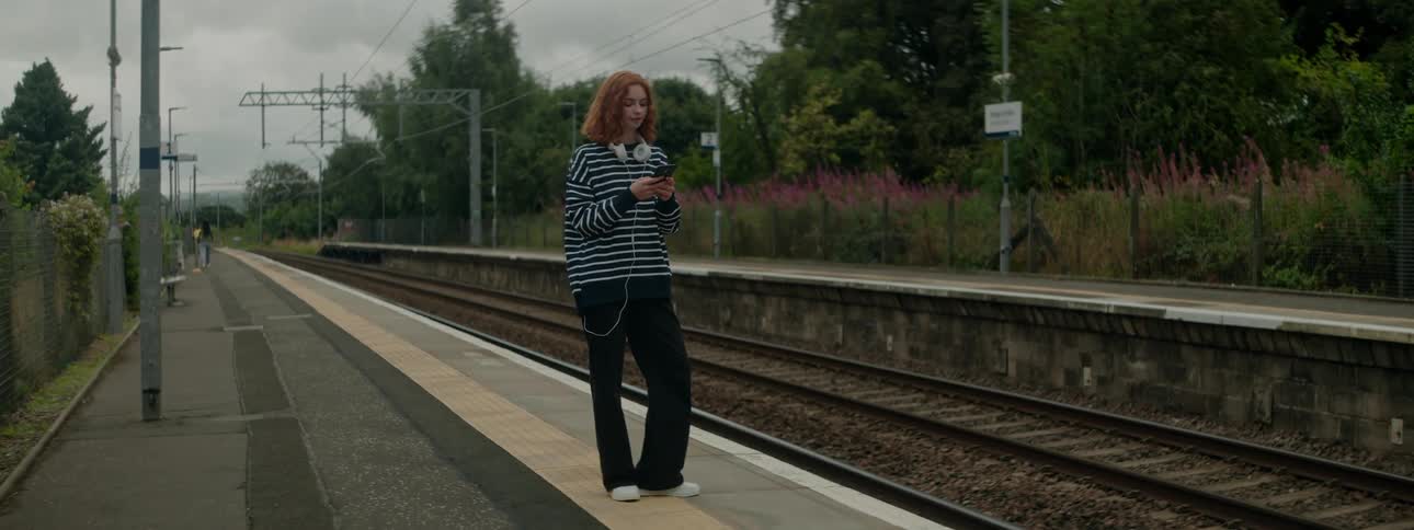 A woman in a striped shirt and with a white headset around her neck is using her cell phone next to a train track