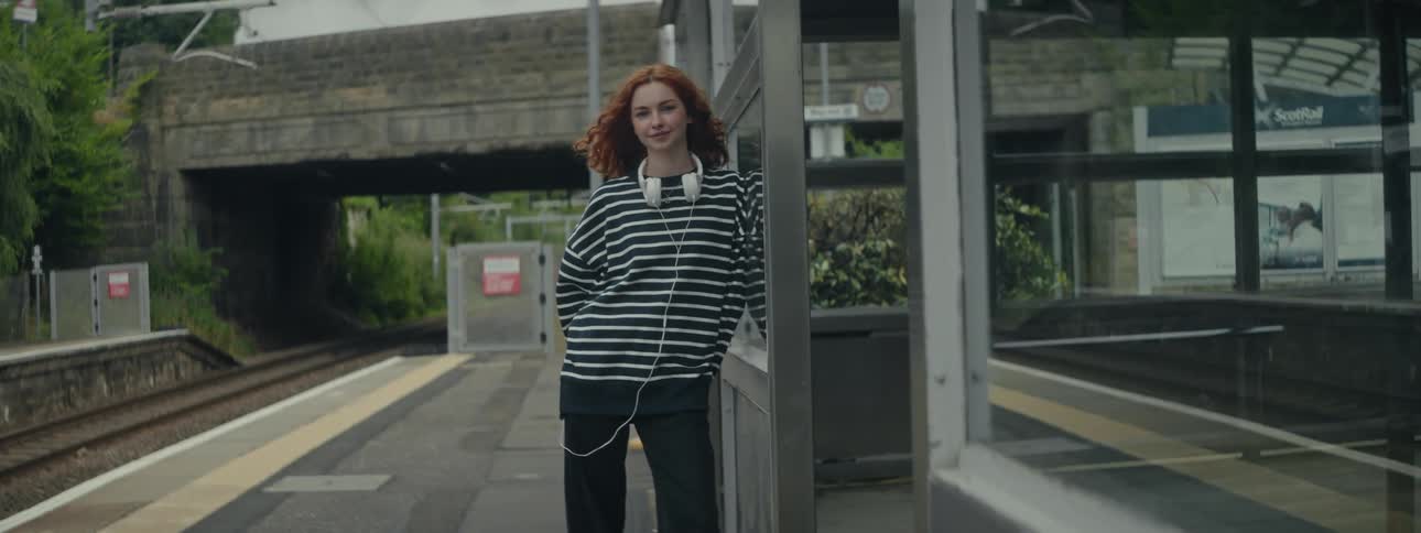 Redhaired woman with white headphones leaning against the side of a booth outside a train terminal