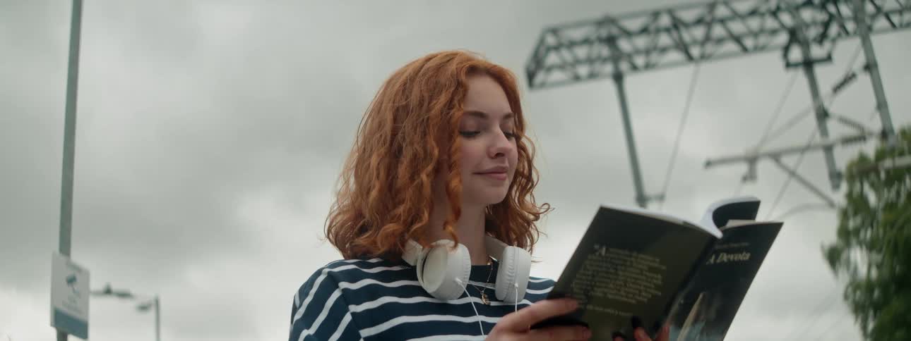 A young woman with red curly hair and white headphones around her neck stands outdoors and reads a book