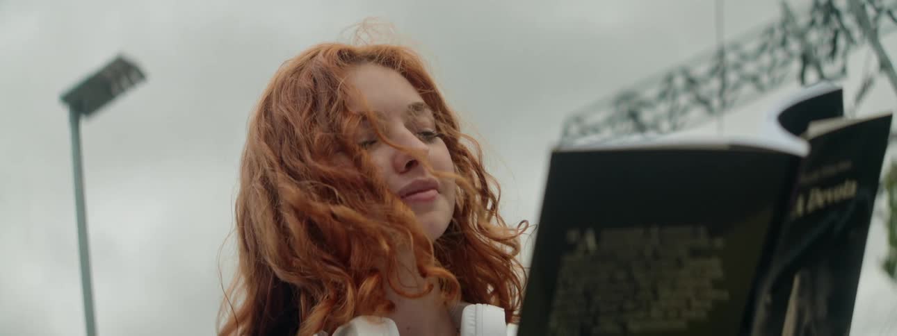 The face of a redhaired woman who is reading a greencovered book held in her hands and she has a headphone around her neck