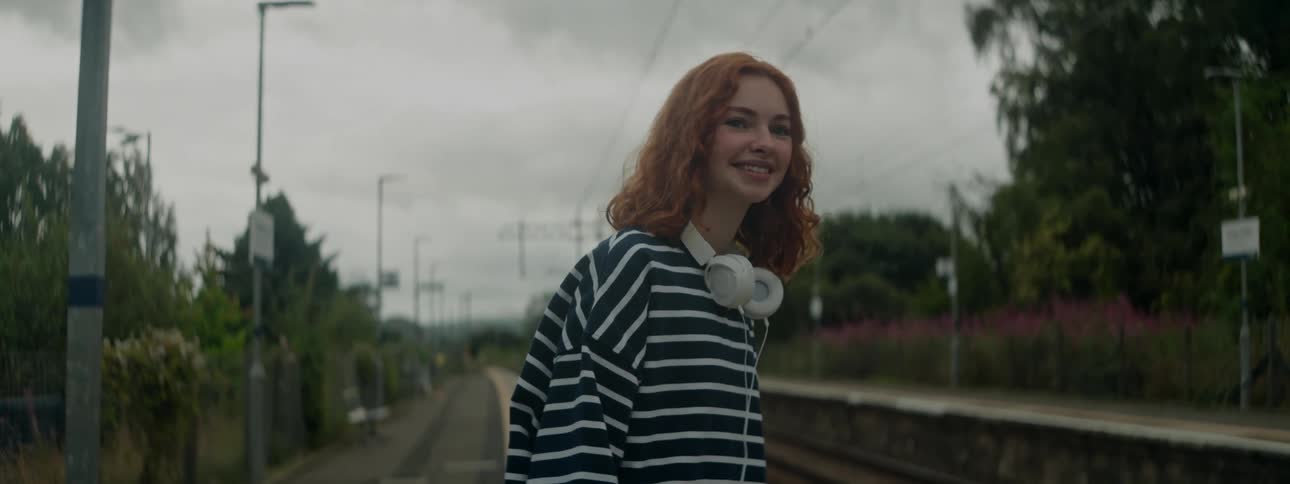 A cheerful woman with red curly hair white headphones and black and white striped sweatshirt stands on a platform
