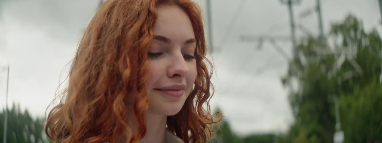 A young woman with long curly red hair smiling slightly as she looks down