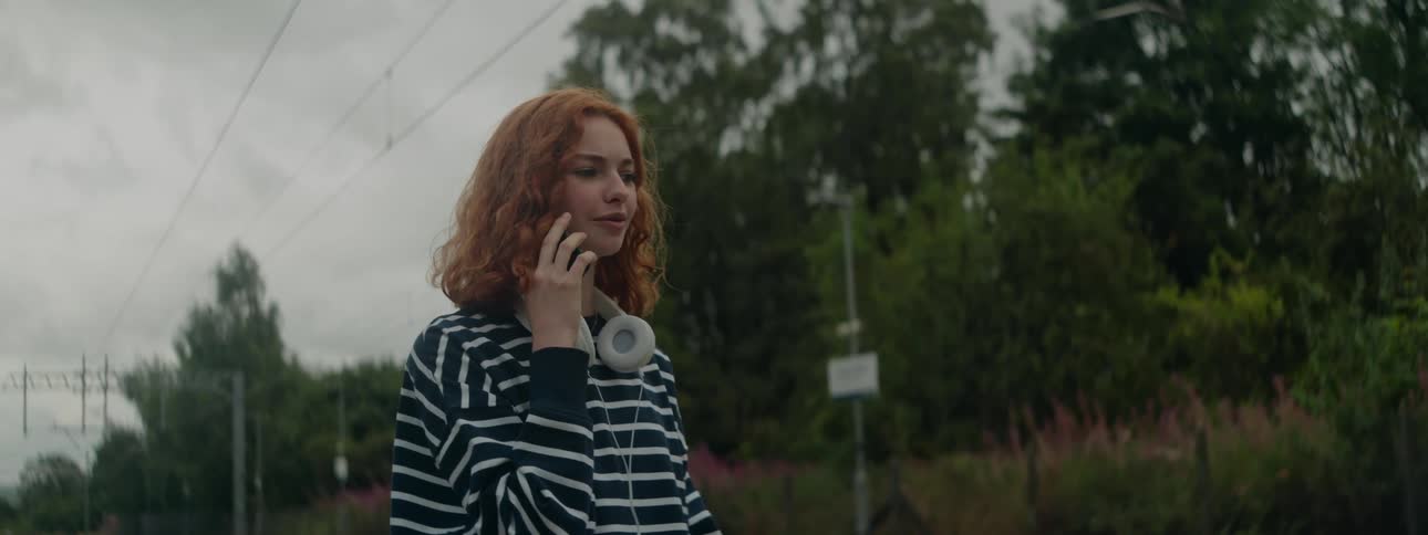 A young woman with curly red hair wearing a striped hoodie and white headphones around her neck smiles while talking on her phone outdoors with lush greenery and train tracks in the background