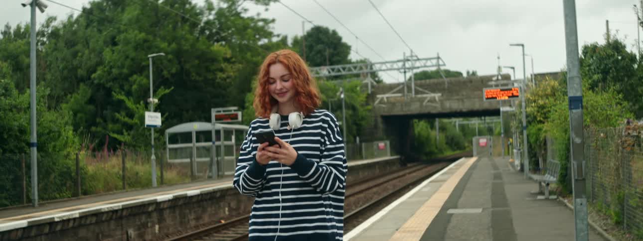 A young redheaded woman is smiling while typing on her cell phone next to a train station