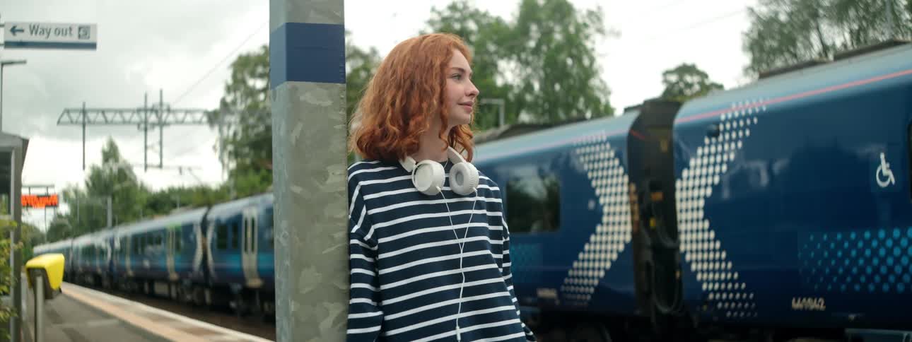 Young redheaded woman at an outdoor station next to a train and leaning against a pole