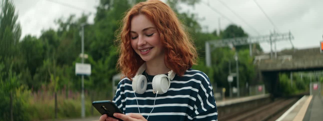 Smiling redhead walks along the railroad tracks while scrolling on her smartphone on an overcast day