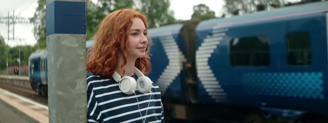 Redheaded woman leaning against a post smiles while the train passes