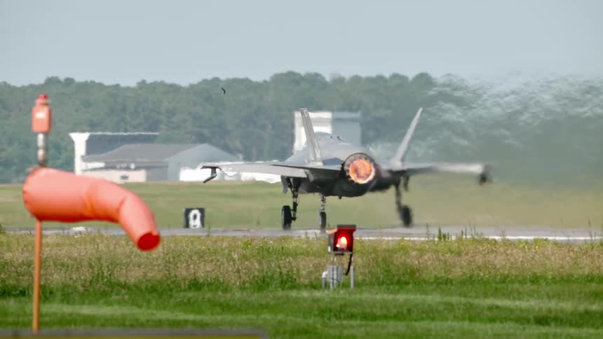 A fighter jet with a glowing afterburner taxis down a runway kicking up heat waves and water vapor