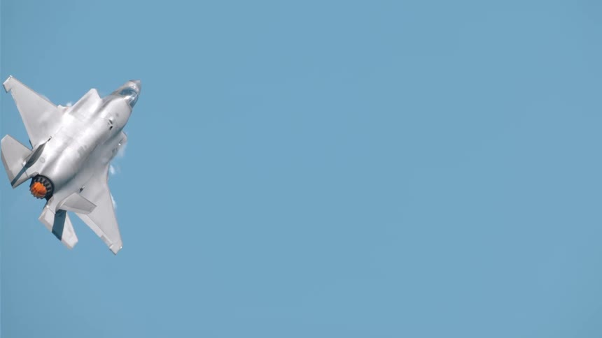 A fighter jet executes a slow tilting maneuver against a clear blue sky