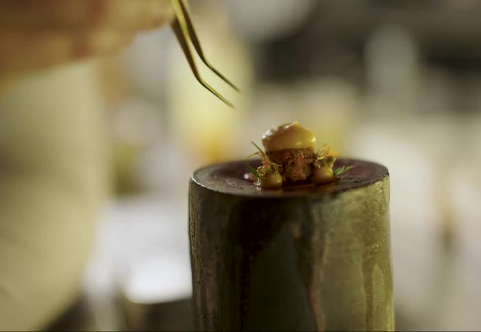 A tiny artfully plated dish featuring a piece of meat sauce and garnish sits atop a cylindrical stand while a chef uses tweezers to make a final adjustment