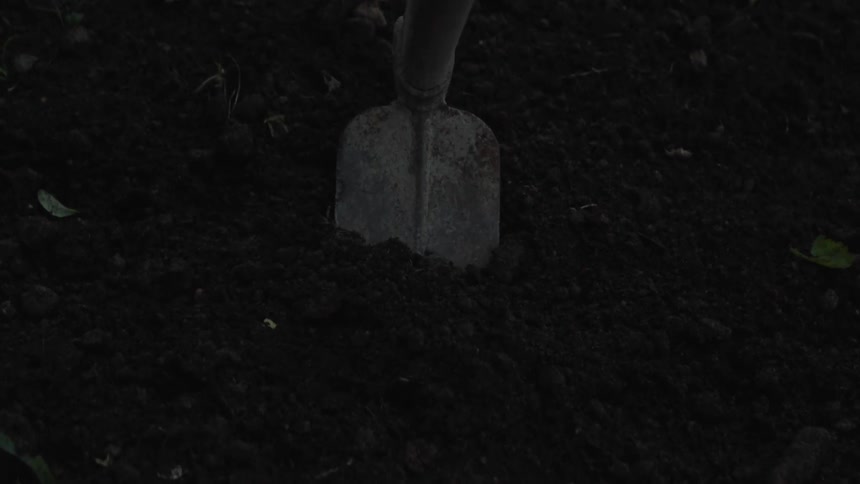 A worn metal shovel blade digging into the ground is shown in dim light creating a dark and moody atmosphere