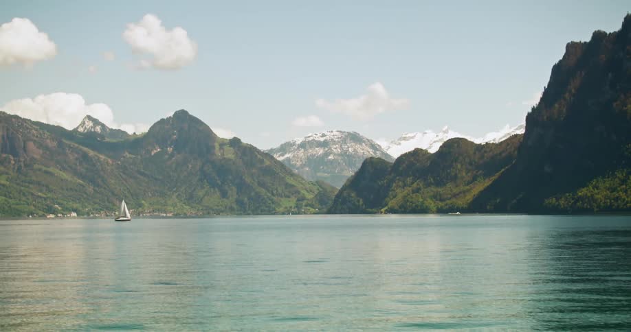 A landscape of a large lake with a small sailboat sailing and behind it there are huge mountains