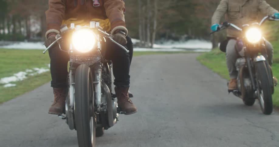 Two men are riding their motorcycles down a road in an area near woods