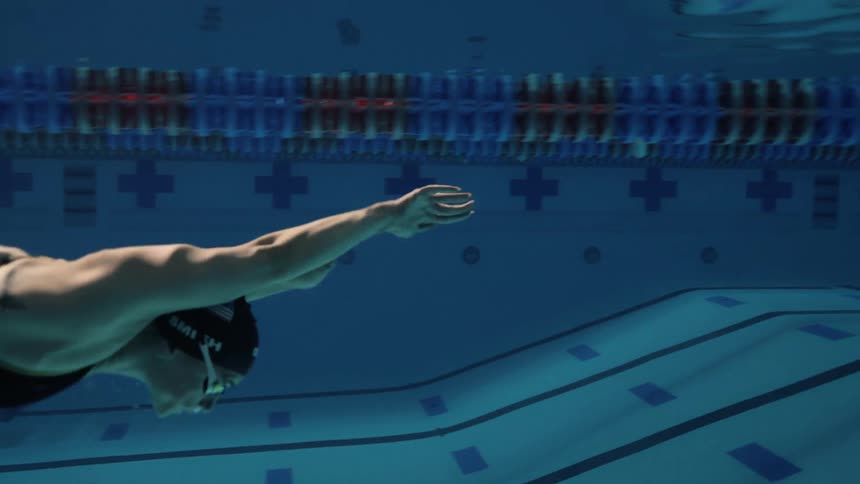 A swimmer is captured midstroke underwater in a pool gliding forward with their arm extended wearing a swim cap and goggles with the pools lane dividers and markings visible in the background