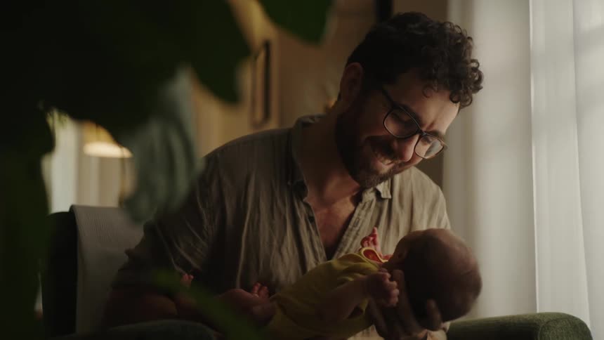 A man with glasses gently holding a baby in his arms gazing at the infant with a tender and loving expression in a softly lit cozy room