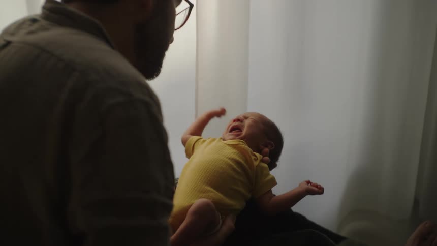 A person is holding a baby dressed in a yellow outfit set against a softly lit background with white curtains