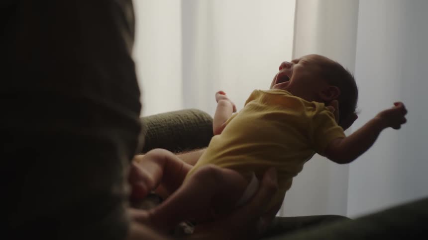 Parent sits next to a curtained window holding a crying infant