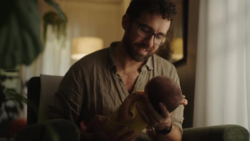 Father smiles at the child he is holding while sitting at home near a window