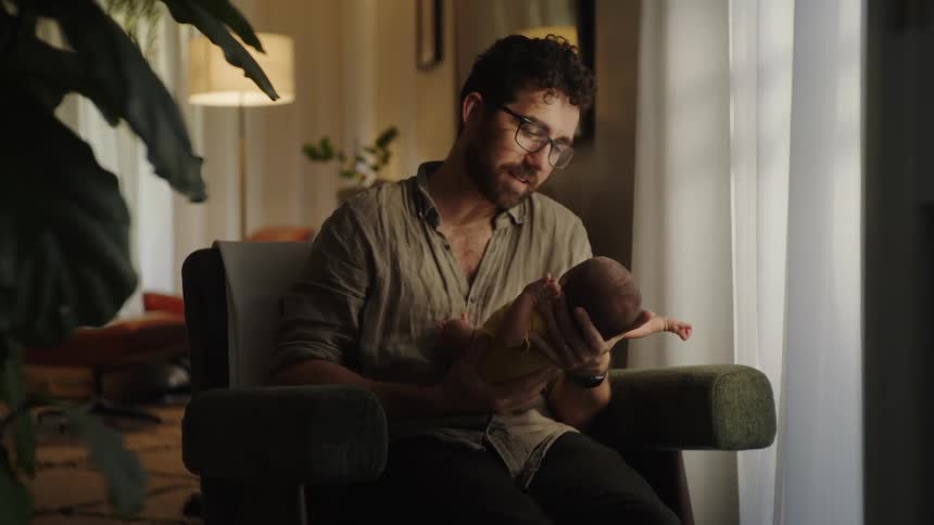 A bearded father with glasses sits in a green armchair holding and talking to a newborn baby