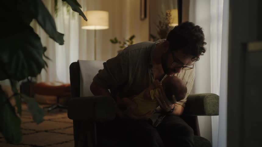 A father with glasses gently holding a newborn baby in his arms gazing at the infant with a tender and loving expression and kissing his forehead in a softly lit cozy room
