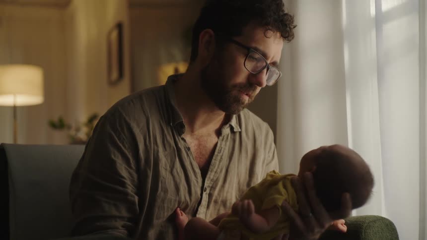 A bearded bespectacled father cradles a newborn baby in a plush green armchair gently talking to the infant