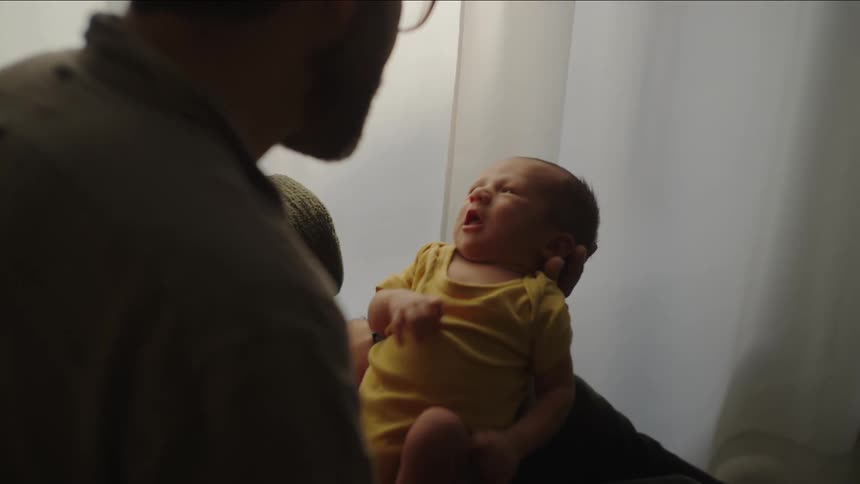 A parent holds their newborn baby who is wearing a yellow onesie while sitting near a window