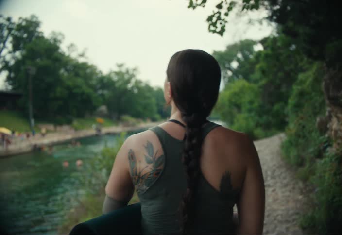 A woman with tattoos on her back and shoulders and her hair in a braid walks away from the camera toward a body of water holding a mat