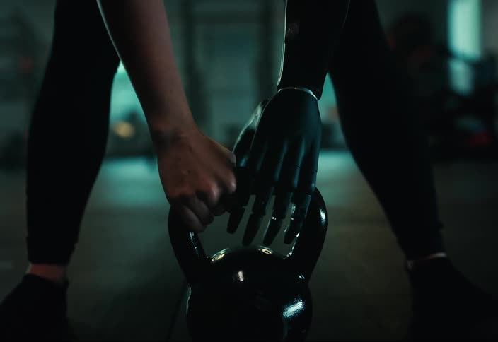 Woman with an artificial limb walks forward to grasp a weight in a gym