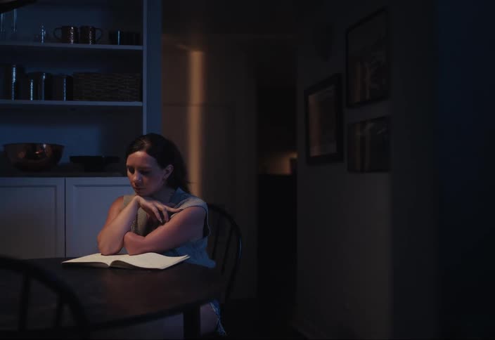 A woman is sitting at a table in a dimly lit room reading an open book at night