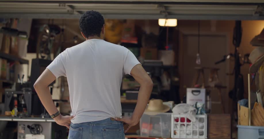 Man wearing a white t shirt and jeans standing in front of a garage filled with various items including boxes a bike and a light hanging from the ceiling