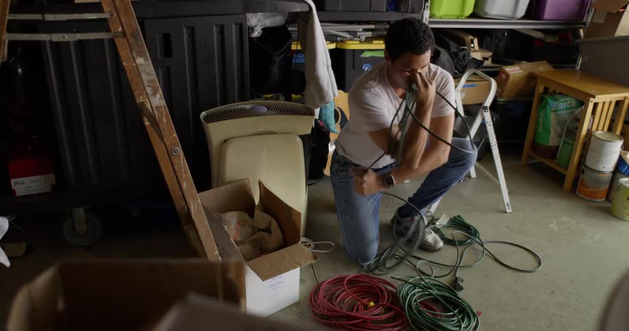 A man in a blue shirt is crouching on the floor of a warehouse and wrapping a green rope around his arm