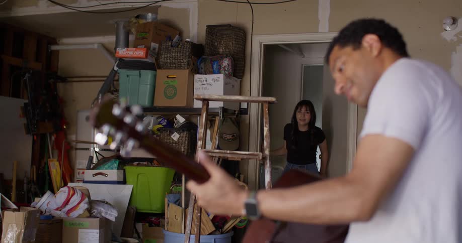 A person is playing a guitar in a cluttered garage filled with various items such as boxes bins and tools while another person is seen entering a room in the background