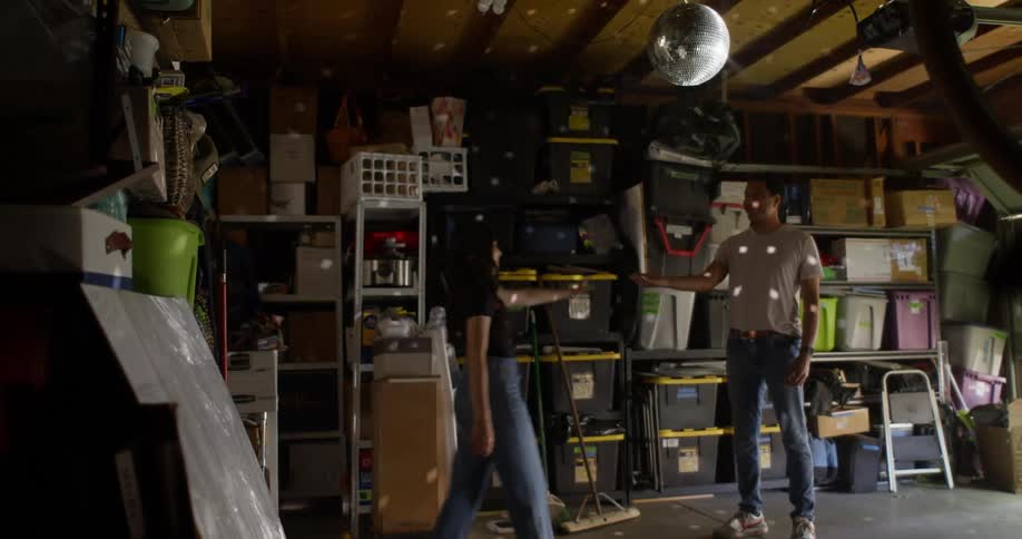 A cluttered garage with shelves full of storage bins and boxes where two people dance under a disco ball hanging from the ceiling