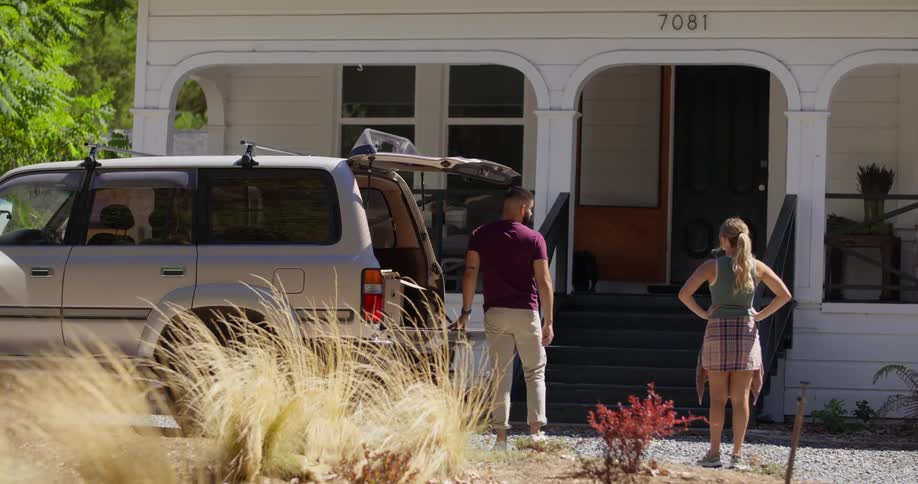 Man opening the trunk of a car and his wife in front of a house and they both hug each other