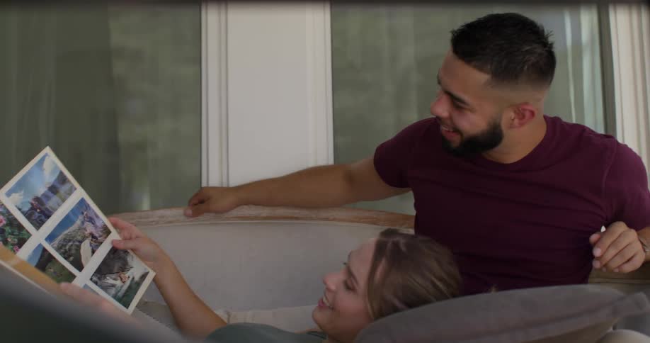 A blonde woman wearing a green top and brown skirt lies on the lap of a bearded man in a red tshirt as she flips through a photo album laughing together