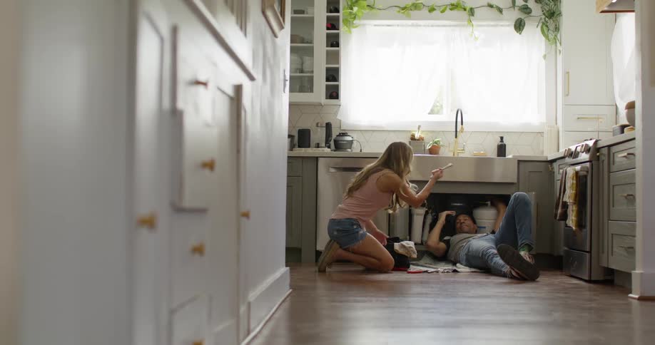 The plumber is repairing the pipes while a blonde woman kneels beside him in the kitchen