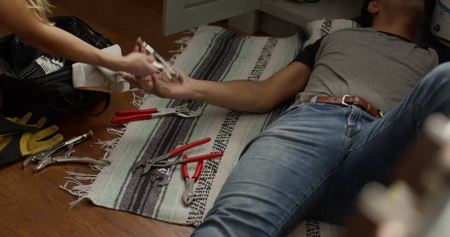 Man lying on the floor picking up tools that are handed to him by another womans hand