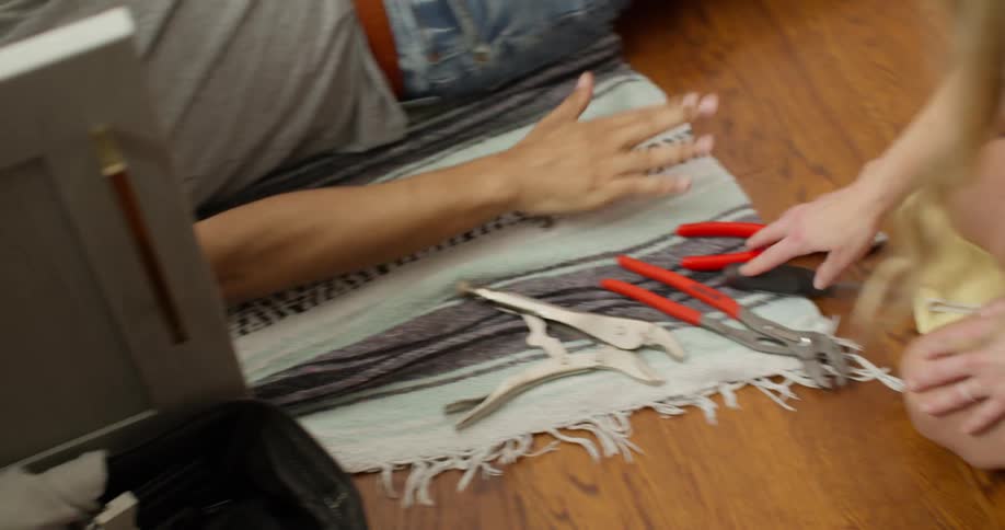 A man lying under a sink fixing it receives a pair of pliers from a woman