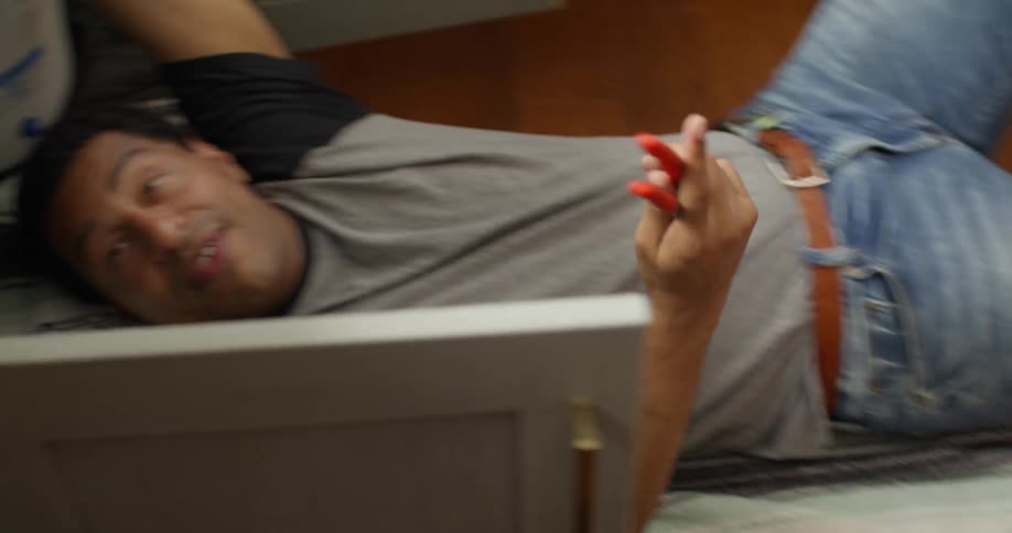 A man wearing a gray shirt with black sleeves is lying on a blanket and is handed a pair of pliers while working on a plumbing project