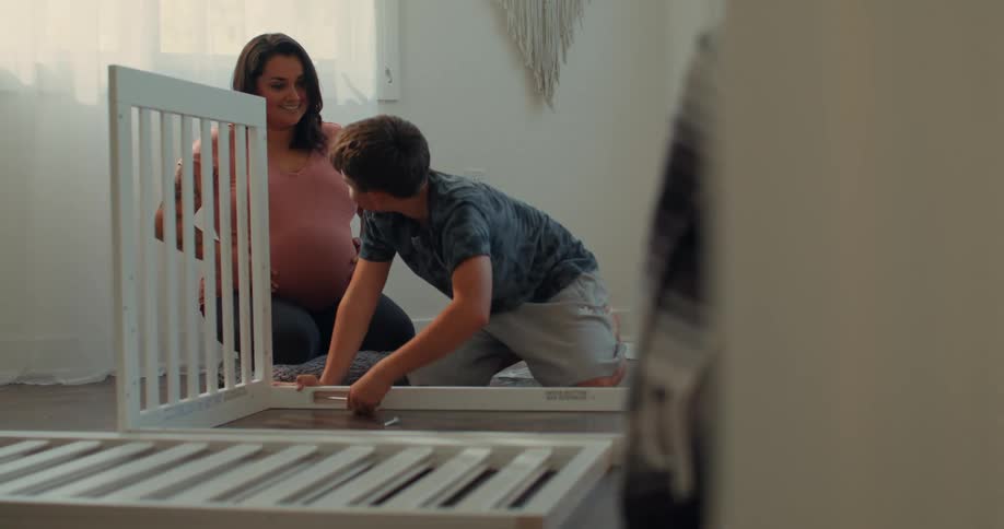 A heavily pregnant mom wearing a pink shirt talks with her son as he assembles a crib in a room