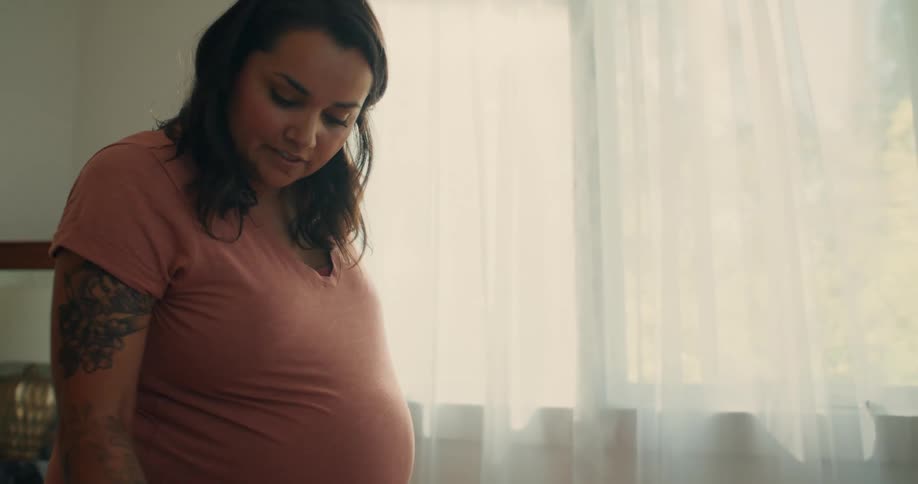 A pregnant woman with her arm tattooed is standing looking at the floor and shaking her head in confirmation tones