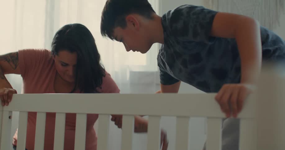 A son helps his pregnant mom with arm tattoos assemble a crib and then helps her up from the ground in the room