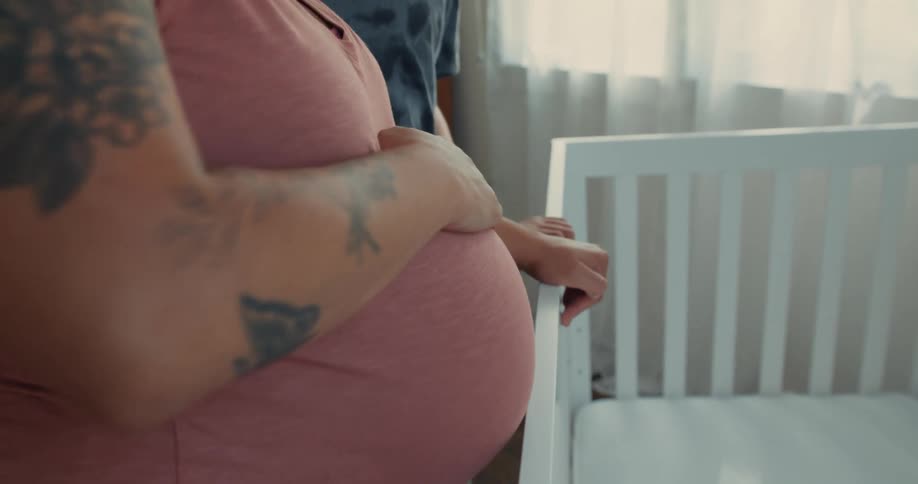 A pregnant woman is rubbing her belly in front of an empty crib