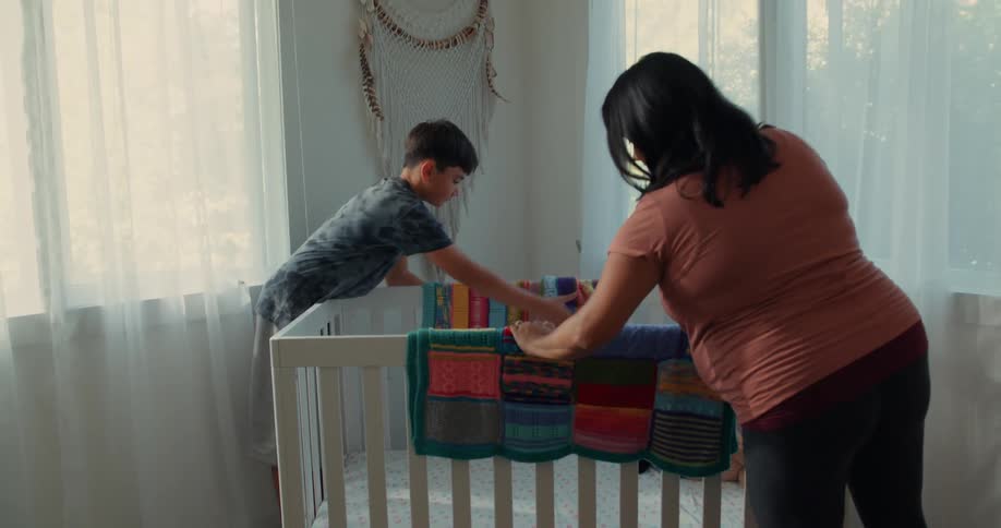 A pregnant woman and a child are gently placing blankets on a crib preparing a cozy space for a baby