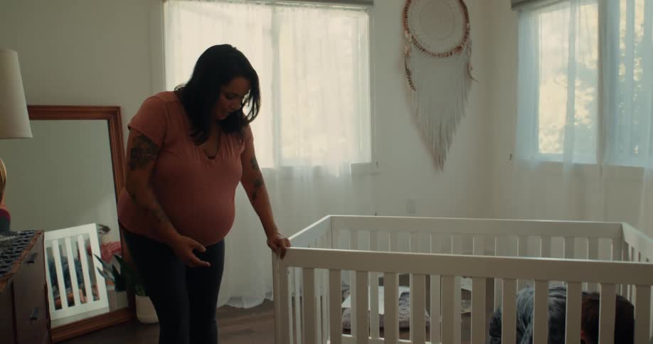 A pregnant woman walking and crouching near a white crib that is being assembled