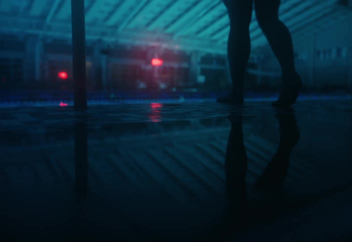 A barefoot woman walks to the edge of an indoor pool in a dimly lit area