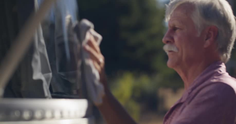 An older man with a mustache wearing a red tshirt and sporting tattoos on his left arm wipes the trailer with a cloth