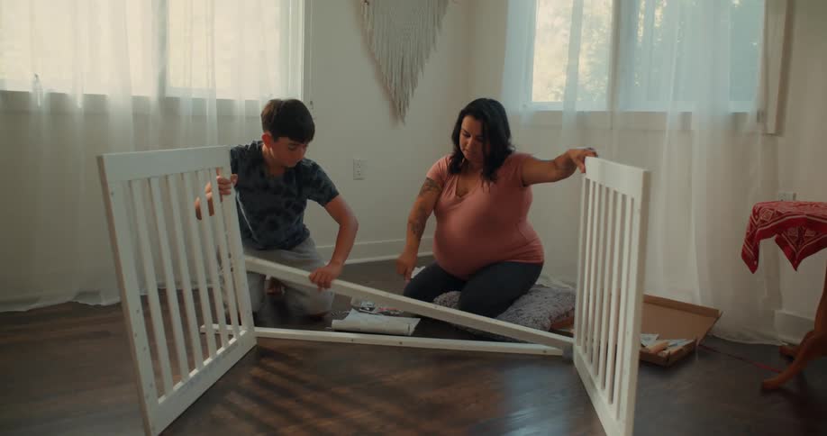 A mother and a child are assembling a white crib in a brightly lit room with wooden floors