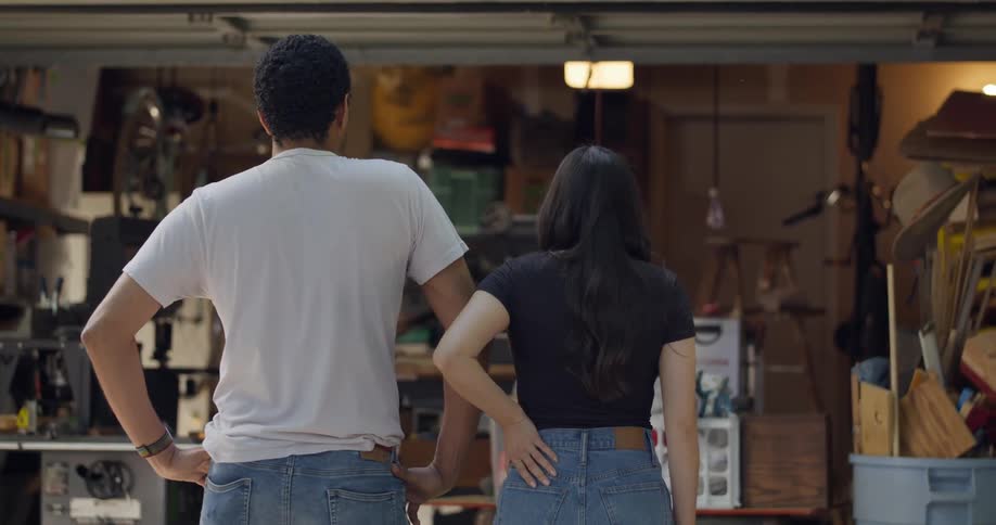 Man and a woman stand in front of a garage with boxes and storage and the woman slaps the man on the back