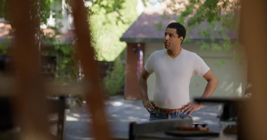 A person wearing a white tshirt and jeans stands outdoors in a driveway with hands on hips looking at a garage